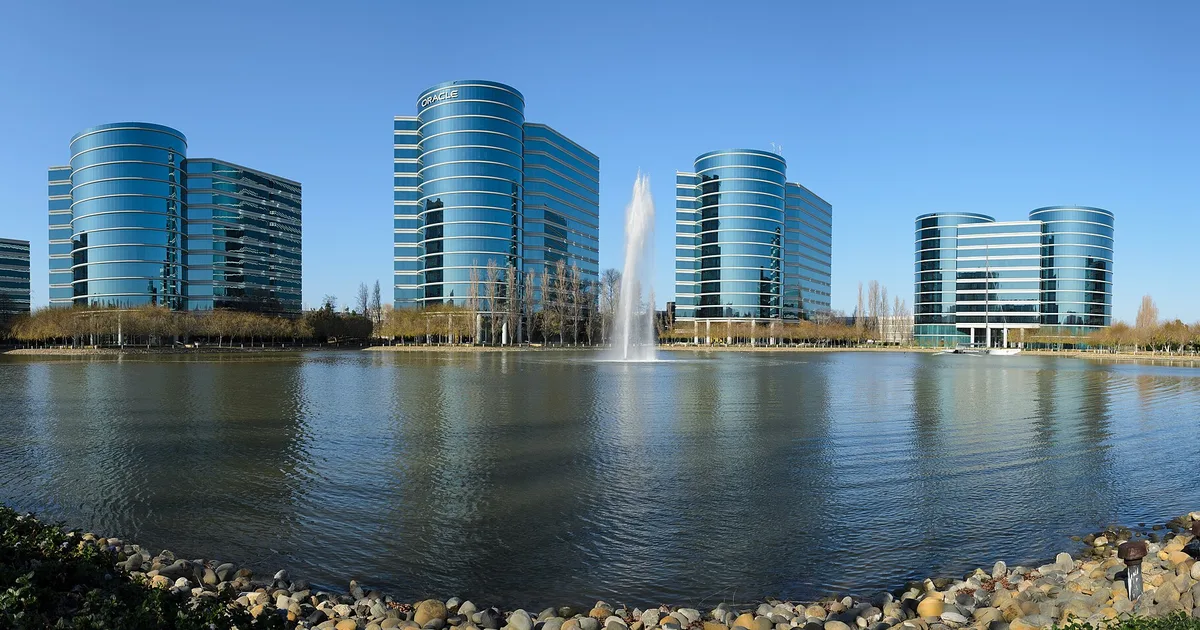 Four curved glass office towers beside an artificial lake with a tall fountain, under a clear blue sky, at Oracle's headquarters in Redwood Shores, California.